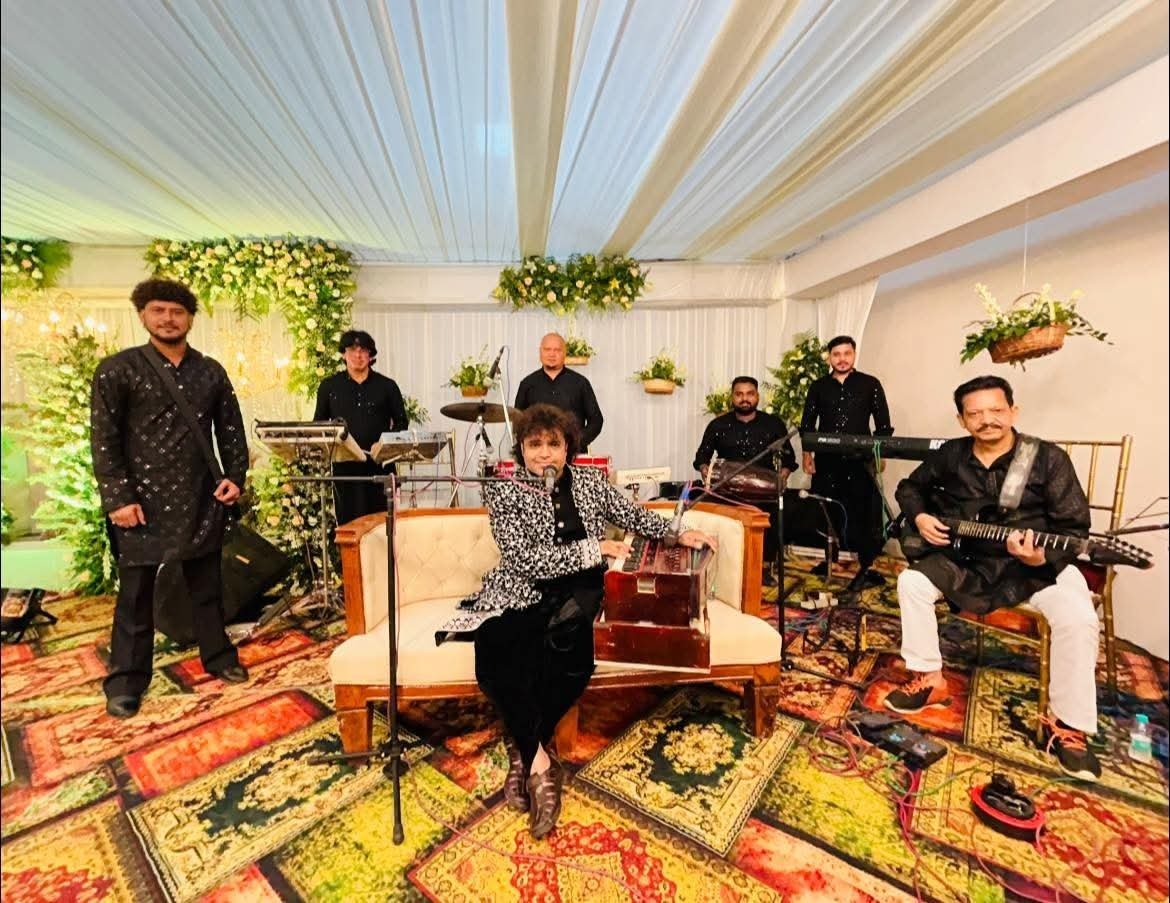 Seven-piece live orchestra with harmonium, tabla, drums and guitar performing at a floral wedding mandap
